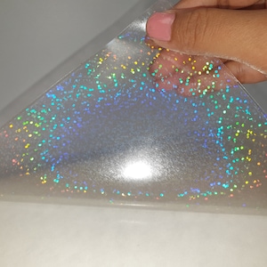 Op de afbeelding: Een transparant zelfklevend vel met een holografisch glitterpatroon dat licht reflecteert in diverse kleuren, waaronder blauw, groen, geel en roze. Het materiaal is flexibel en helder, met een dichte verdeling van kleine, glinsterende stippen.