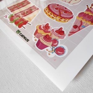 May include: A sheet of stickers featuring various pink and red desserts, including cupcakes, cake slices, ice cream cones, and candy apples. The stickers are on a white background with a pink and white grid pattern. The text "CATKUS" is visible in the bottom left corner.