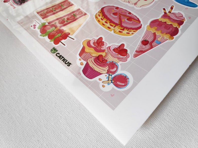 May include: A sheet of stickers featuring various pink and red desserts, including cupcakes, cake slices, ice cream cones, and candy apples. The stickers are on a white background with a pink and white grid pattern. The text "CATKUS" is visible in the bottom left corner.