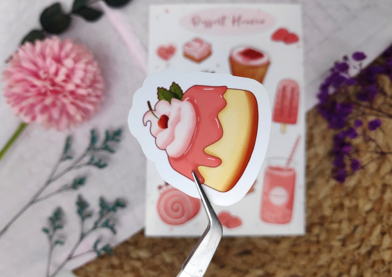 May include: A pink and yellow sticker with a cartoon illustration of a cake with pink frosting and a cherry on top. The sticker is part of a set of stickers with a "Sweet House" theme.