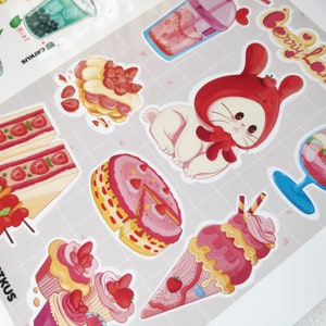 May include: A set of colourful stickers featuring a variety of sweet treats, including cakes, ice cream, and a bunny wearing a strawberry hat. The stickers are decorated with strawberries and hearts and have the text "Berry Land!" on one of the stickers.