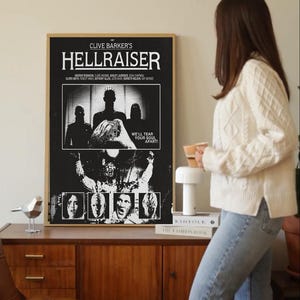 Może przedstawiać: Oprawiony czarno-biały plakat filmowy do filmu "Hellraiser" Clive'a Barkera. Plakat zawiera tytuł filmu, obsadę i graficzny obraz. Plakat znajduje się na drewnianej szafce.