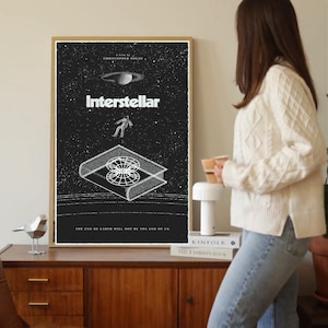 May include: A framed black and white poster for the film "Interstellar" in a gold frame. The poster features the film's title, an astronaut, a planet, and a book-like structure. The text "The End of Earth Will Not Be the End of Us" is at the bottom.
