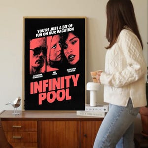May include: A framed movie poster for "Infinity Pool" with a black background and red text. The poster features the faces of Alexander Skarsgard, Mia Goth, and Cleopatra Coleman. The text "YOU'RE JUST A BIT OF FUN ON OUR VACATION" is at the top.