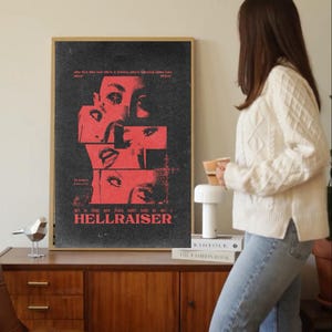 Puede incluir: Un póster enmarcado con fondo negro y elementos gráficos rojos. El póster presenta un collage de rasgos faciales y el título "HELLRAISER" en texto rojo. El póster se exhibe en un mueble de madera.