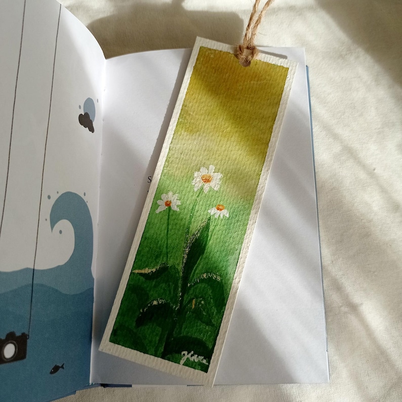 May include: Watercolor bookmark featuring a field of white daisies with yellow centers and green leaves against a gradient yellow and green background. The bookmark is rectangular with a string attached at the top.