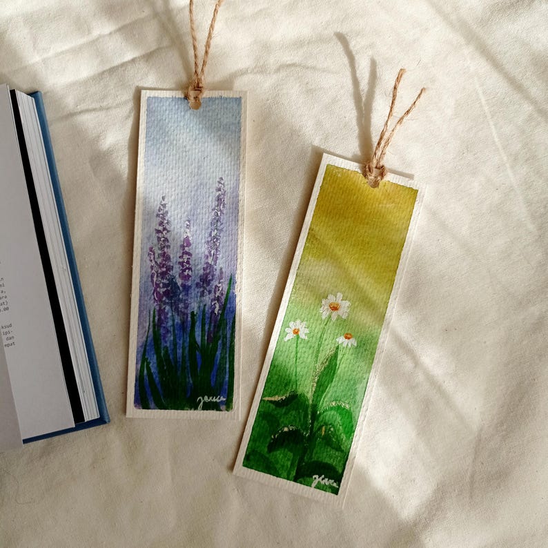 May include: Two watercolor bookmarks with twine ties. One features lavender, the other daisies. The lavender bookmark has a blue and purple background with green grass. The daisy bookmark has a yellow and green background. Both have the artist's signature.