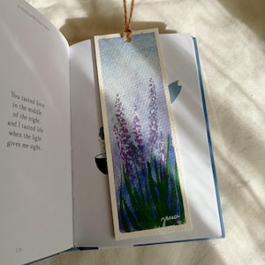 May include: Watercolor bookmark featuring lavender flowers with purple blooms and green stems against a light blue sky. The bookmark is made of textured paper and has a brown string attached. The bookmark is placed in an open book.