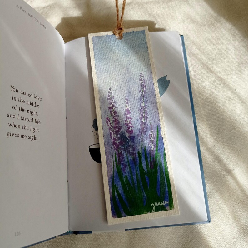 May include: Watercolor bookmark featuring lavender flowers with purple blooms and green stems against a light blue sky. The bookmark is made of textured paper and has a brown string attached. The bookmark is placed in an open book.
