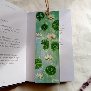 May include: A rectangular bookmark featuring a watercolor painting of a pond scene with lily pads and white water lilies. The bookmark is attached to a book with a brown string. The text "A Poem with Your Name" is at the top.