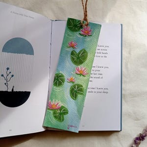 May include: Watercolor bookmark featuring pink water lilies and green lily pads on a blue and green background. The bookmark has a jute string for hanging and the text "A Poem with Your Name" is visible. The artist's signature "Janice" is at the bottom.