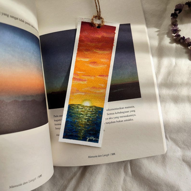 May include: A watercolor bookmark featuring a sunset scene. The bookmark depicts a vibrant sky transitioning from red to orange and yellow, with a blue ocean below. It is attached to a book with text in a foreign language.