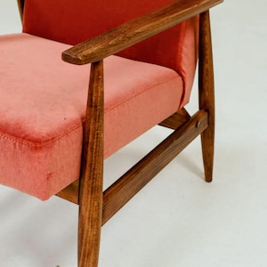 May include: Mid-century modern armchair with a coral-colored seat and backrest. The wooden frame and armrests are a dark brown. The chair has a classic, minimalist design, perfect for living room or bedroom decor.