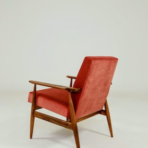 May include: Mid-century modern armchair with a coral-colored velvet seat and backrest. The chair features a wooden frame with armrests and tapered legs. The design is a classic example of mid-century furniture.