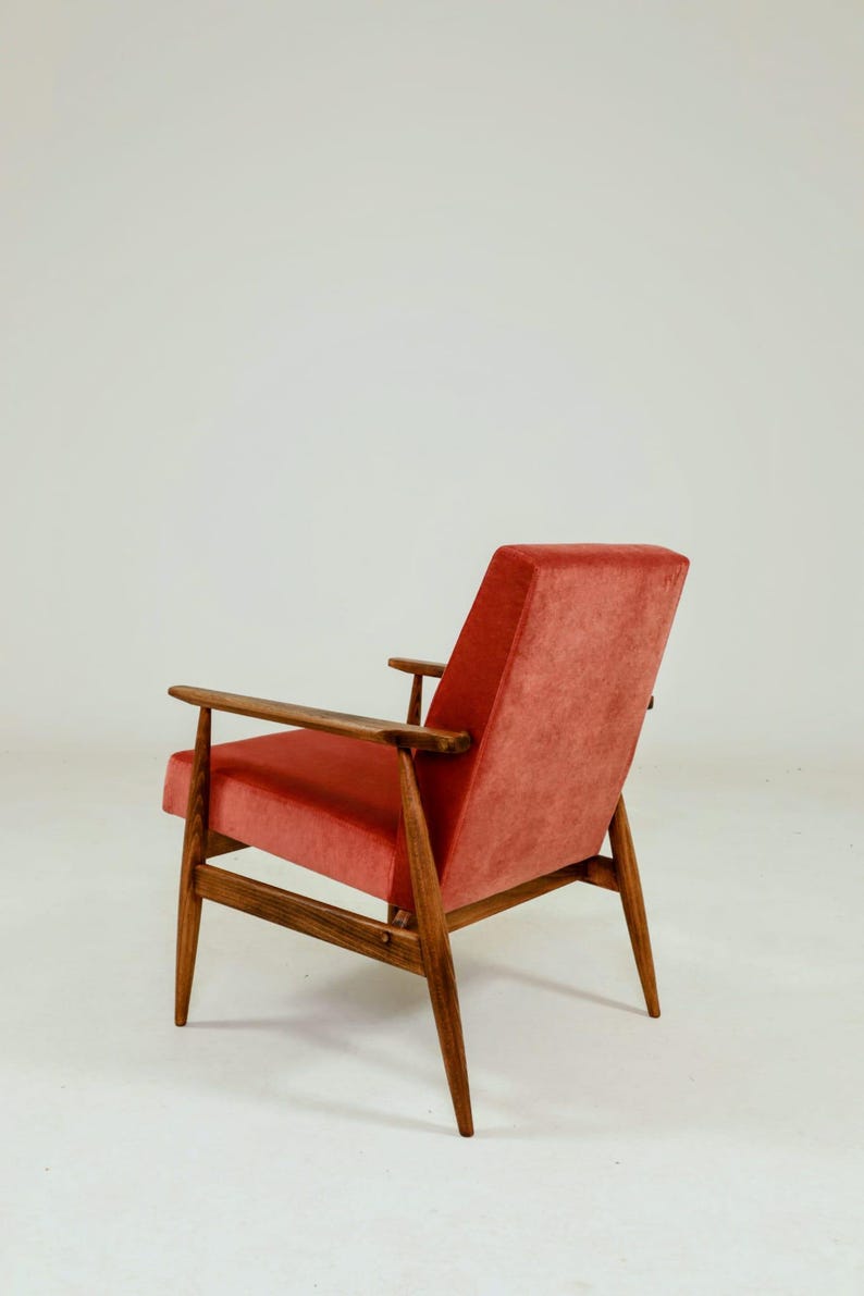May include: Mid-century modern armchair with a coral-colored velvet seat and backrest. The chair features a wooden frame with armrests and tapered legs. The design is a classic example of mid-century furniture.