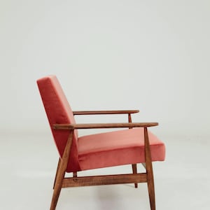 May include: A mid-century modern armchair with a coral-colored velvet seat and backrest. The chair features a wooden frame with armrests and angled legs. The design is simple and elegant, suitable for a living room or lounge area.