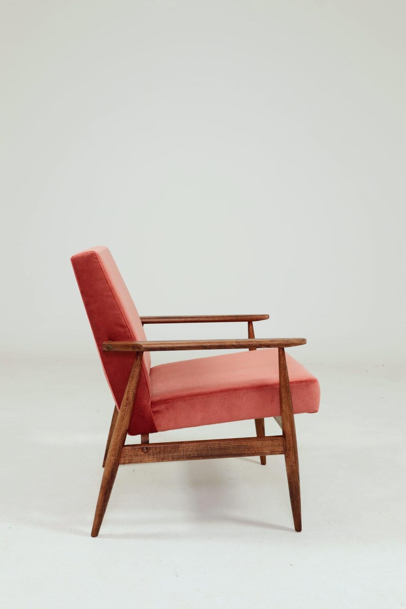 May include: A mid-century modern armchair with a coral-colored velvet seat and backrest. The chair features a wooden frame with armrests and angled legs. The design is simple and elegant, suitable for a living room or lounge area.