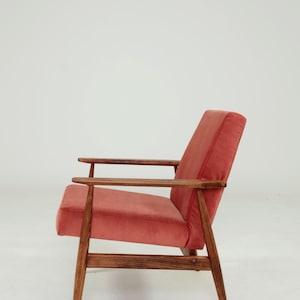 May include: A mid-century modern armchair with a coral-colored velvet seat and backrest. The chair features a wooden frame and armrests, showcasing a minimalist design. The legs are angled, and the overall aesthetic is vintage.