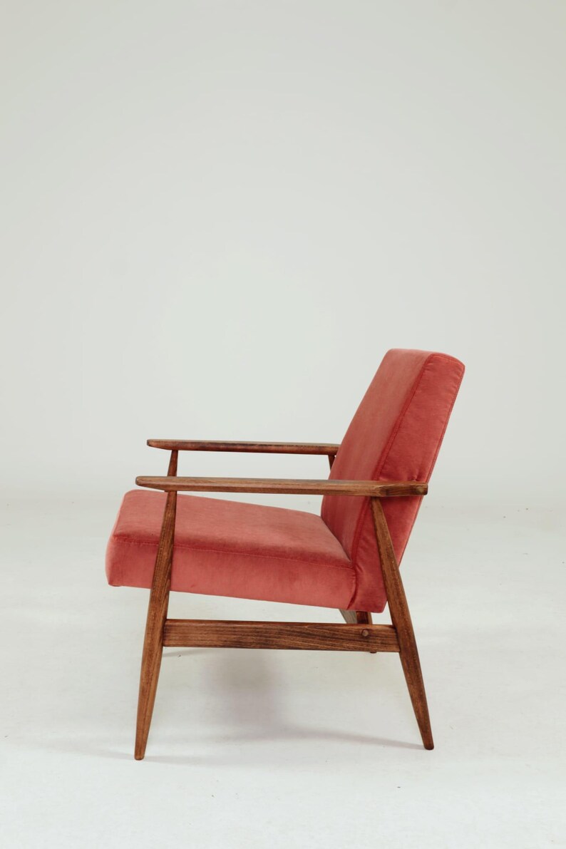 May include: A mid-century modern armchair with a coral-colored velvet seat and backrest. The chair features a wooden frame and armrests, showcasing a minimalist design. The legs are angled, and the overall aesthetic is vintage.