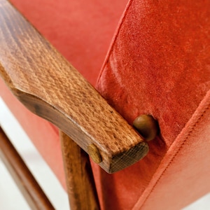 May include: Close-up of a mid-century modern chair with a vibrant red seat and backrest. The chair features wooden armrests and legs, showcasing a warm, natural wood grain. The design highlights the chair's clean lines and retro aesthetic.