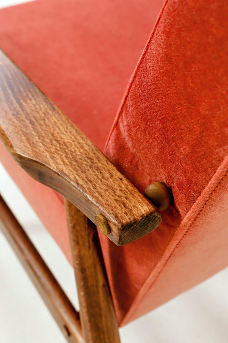 May include: Close-up of a mid-century modern chair with a vibrant red seat and backrest. The chair features wooden armrests and legs, showcasing a warm, natural wood grain. The design highlights the chair's clean lines and retro aesthetic.