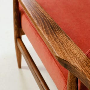 May include: Close-up of a mid-century modern armchair. The chair features a wooden frame with a rich, warm brown finish. The seat and backrest are upholstered in a vibrant, coral-colored fabric. The armrest is made of wood.