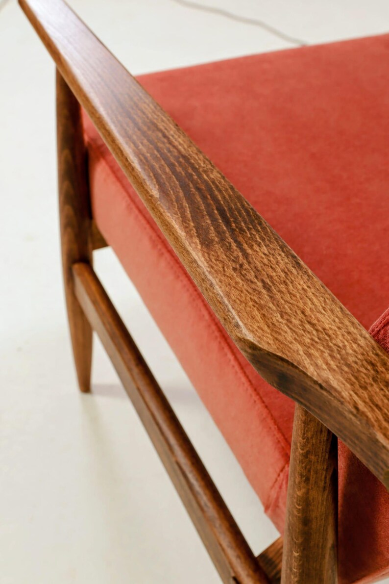 May include: Close-up of a mid-century modern armchair. The chair features a wooden frame with a rich, warm brown finish. The seat and backrest are upholstered in a vibrant, coral-colored fabric. The armrest is made of wood.