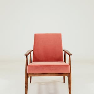 May include: A mid-century modern armchair with a coral-colored velvet seat and back. The chair has a wooden frame with angled legs and armrests. The design is simple and elegant, suitable for a living room or lounge area.