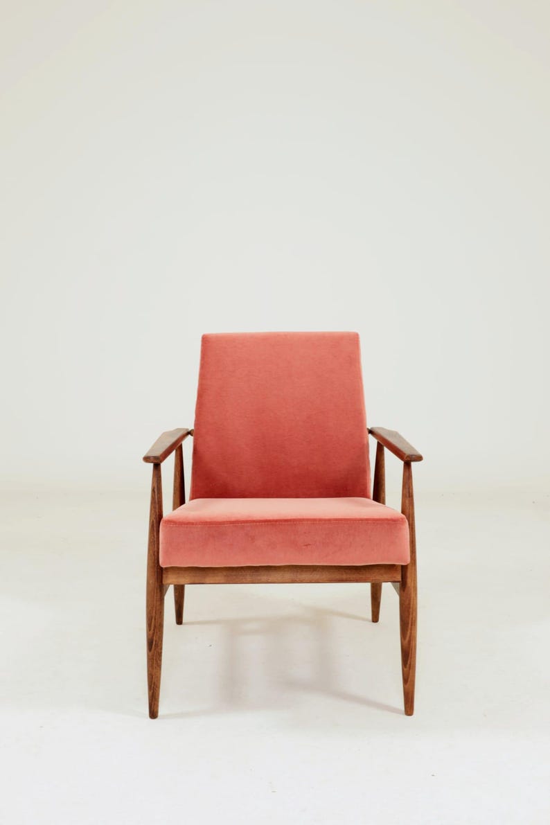 May include: A mid-century modern armchair with a coral-colored velvet seat and back. The chair has a wooden frame with angled legs and armrests. The design is simple and elegant, suitable for a living room or lounge area.