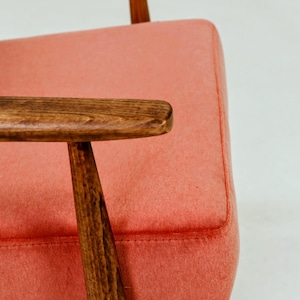 May include: Close-up of a mid-century modern armchair. The chair features a coral-colored upholstered seat and backrest, with wooden armrests and legs. The wood has a warm, brown tone, and the design is simple and elegant.