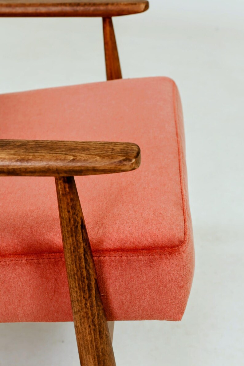 May include: Close-up of a mid-century modern armchair. The chair features a coral-colored upholstered seat and backrest, with wooden armrests and legs. The wood has a warm, brown tone, and the design is simple and elegant.