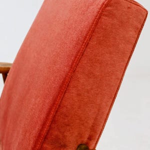 May include: Close-up of a mid-century modern chair. The chair features a vibrant, reddish-orange upholstered back and seat. The wooden armrest is a warm, medium brown. The stitching on the back is visible.