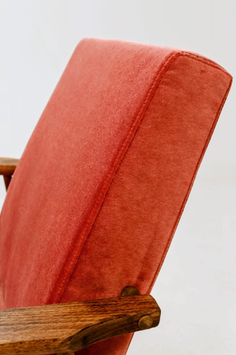 May include: Close-up of a mid-century modern chair. The chair features a vibrant, reddish-orange upholstered back and seat. The wooden armrest is a warm, medium brown. The stitching on the back is visible.