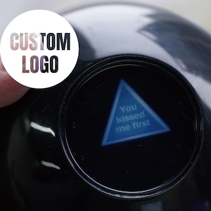 May include: A black spherical fortune-telling toy featuring a circular white sticker with the text CUSTOM LOGO. Inside the clear window, a blue triangular prism displays the white text You kissed me first against a dark background.