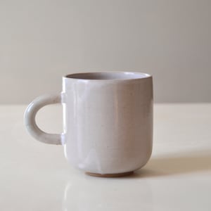 Puede incluir: Una taza de cerámica blanca con un asa redondeada. La taza tiene una superficie ligeramente texturizada y una base redondeada.
