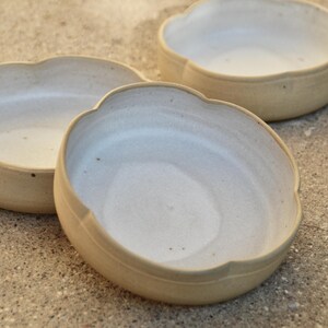 May include: Three white ceramic bowls with scalloped edges. The bowls are arranged in a staggered formation on a light brown surface.