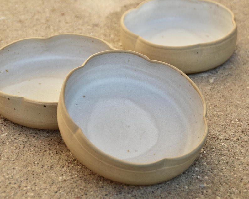 May include: Three white ceramic bowls with scalloped edges. The bowls are arranged in a staggered formation on a light brown surface.