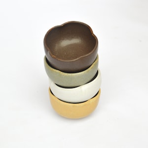 Op de afbeelding: Een stapel van vier keramische kommen in verschillende kleuren. De bovenste kom is bruin, de tweede is groen, de derde is wit en de onderste kom is geel.