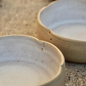 May include: Two white ceramic bowls with a slightly textured surface. The bowls have a rounded shape with a slightly flared rim.