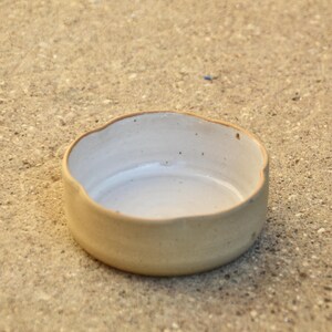 May include: A small, white ceramic bowl with a slightly irregular rim. The bowl is sitting on a light brown surface.
