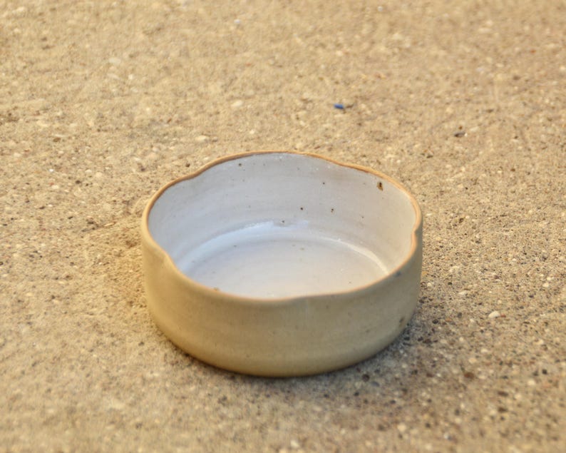 May include: A small, white ceramic bowl with a slightly irregular rim. The bowl is sitting on a light brown surface.