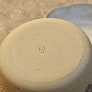 May include: A beige ceramic bowl with a rough texture and a small, stamped mark on the bottom that reads "Icil."