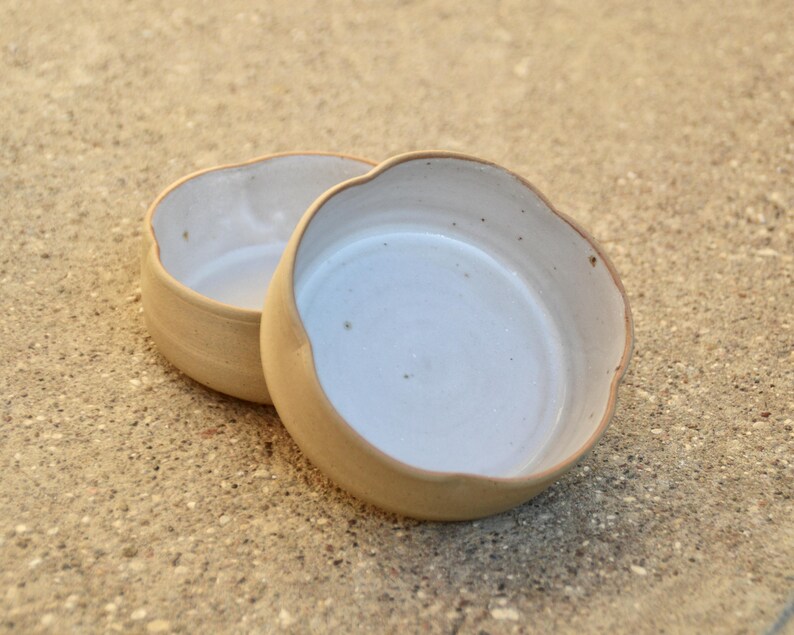 May include: Two ceramic bowls with a white glaze and a light brown rim. The bowls are slightly scalloped and have a rustic, handmade look.