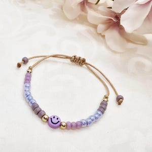 Könnte beinhalten: Verstellbares Armband mit einer lila Smiley-Gesichtsperle und einer Mischung aus lila, lavendelfarbenen und blauen Perlen. Das Armband hat goldfarbene Akzente und eine beige Kordel. Ein lustiges und farbenfrohes Accessoire.