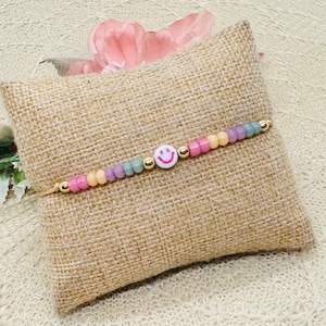Könnte beinhalten: Ein goldenes Kettenarmband mit pastellrosa, gelben und grünen Perlen und einem weißen Smiley-Anhänger.