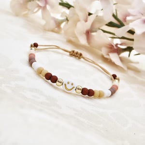 Könnte beinhalten: Armband mit Smiley-Anhänger. Das Armband hat weiße, braune, beige und goldfarbene Perlen. Es ist an einem beigefarbenen Kordelband befestigt und hat einen Schiebeknotenverschluss.
