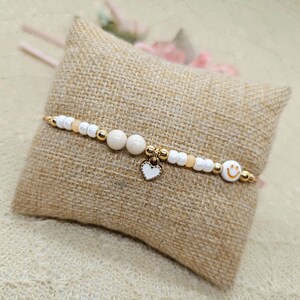 Könnte beinhalten: Ein weißes und pfirsichfarbenes Perlenarmband mit einem goldenen Herz-Anhänger und einem goldenen Smiley-Anhänger.