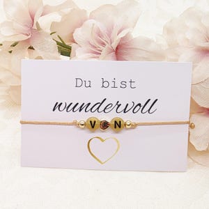 Könnte beinhalten: Ein goldfarbenes Perlenarmband mit den Buchstaben "V", "N" und einer herzförmigen Perle. Das Armband liegt auf einer weißen Karte mit dem deutschen Text "Du bist wundervoll" und einer goldenen Herzgrafik. Rosa Blumen sind im Hintergrund.