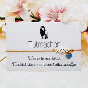 Könnte beinhalten: Goldfarbenes Armband mit einem weißen Smiley-Gesicht-Perle, goldenen Perlen und einem blauen Herz-Anhänger. Das Armband wird auf einer weißen Karte mit dem Wort "Mutmacher" und inspirierendem Text präsentiert.