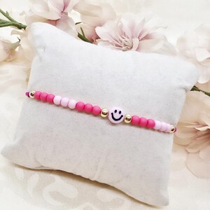 Könnte beinhalten: Ein Perlenarmband mit einer zentralen hellrosa Smiley-Perle, flankiert von metallisch goldenen Perlen und abwechselnd matten, kräftig pinken und hellrosa runden Perlen, die in einem symmetrischen Muster entlang des Armbands angeordnet sind.